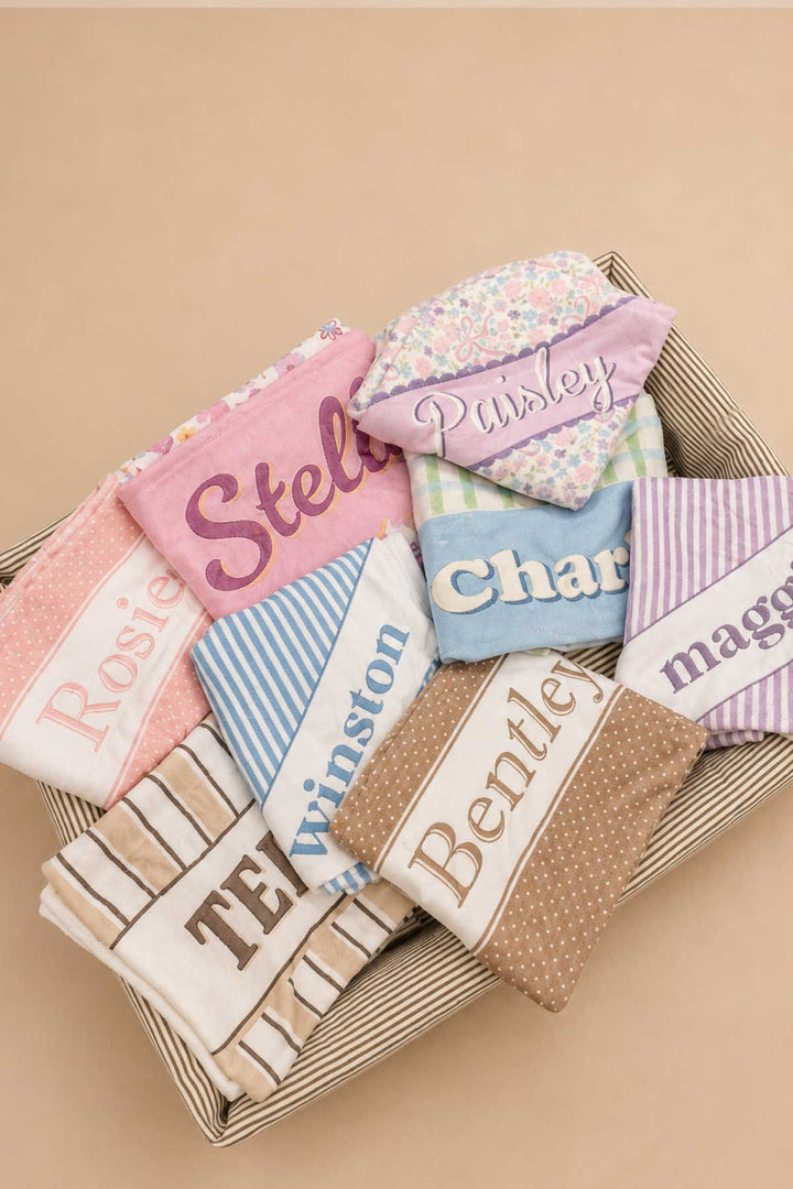 A basket filled with multiple personalized pet blankets in various prints and colors, including the Ribbon Garden print (labeled "Paisley"), showcasing the full range of customization options available