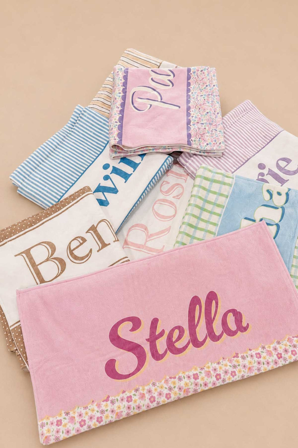 Group shot of multiple personalized pet towels in various prints and colors stacked together, with the front towel showing the name Stella in large pink script lettering