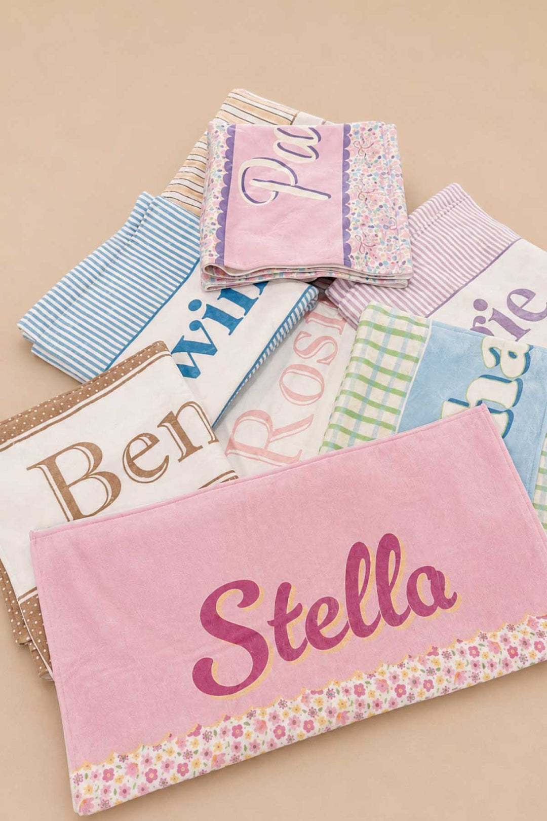Group shot of multiple personalized pet towels in various prints and colors stacked together, with the front towel showing the name Stella in large pink script lettering