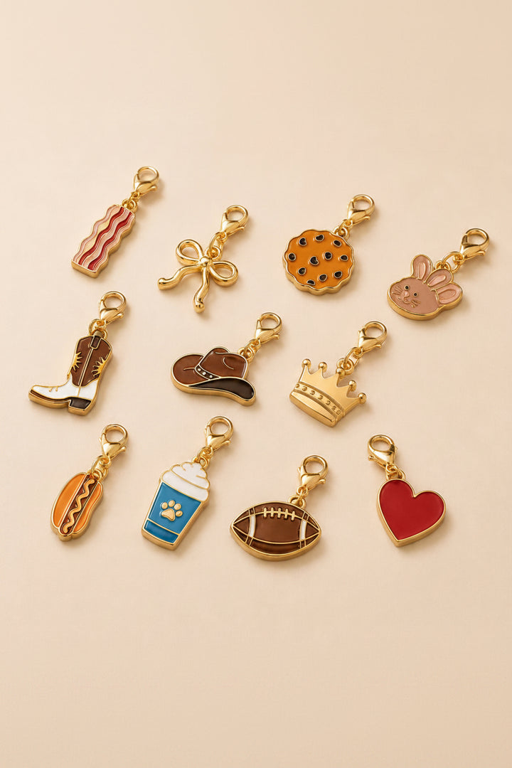 Collection of gold enamel pet collar charms on a blush background, including bacon, bow, cookie, bunny, cowboy boot, cowboy hat, crown, hot dog, pup cup, football, and red heart charms with lobster claw clasps