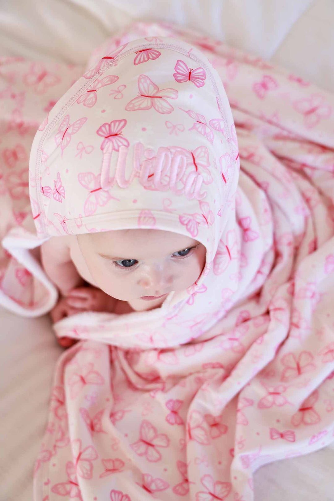 Embroidered Bamboo Hooded Towel in Blooming Butterflies print - baby lying flat wrapped in the pink butterfly-print bamboo towel with embroidered name on the hood, photographed from above