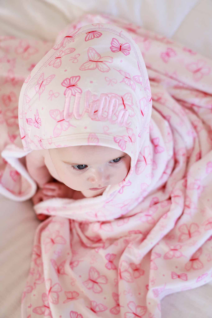 Embroidered Bamboo Hooded Towel in Blooming Butterflies print - baby lying flat wrapped in the pink butterfly-print bamboo towel with embroidered name on the hood, photographed from above