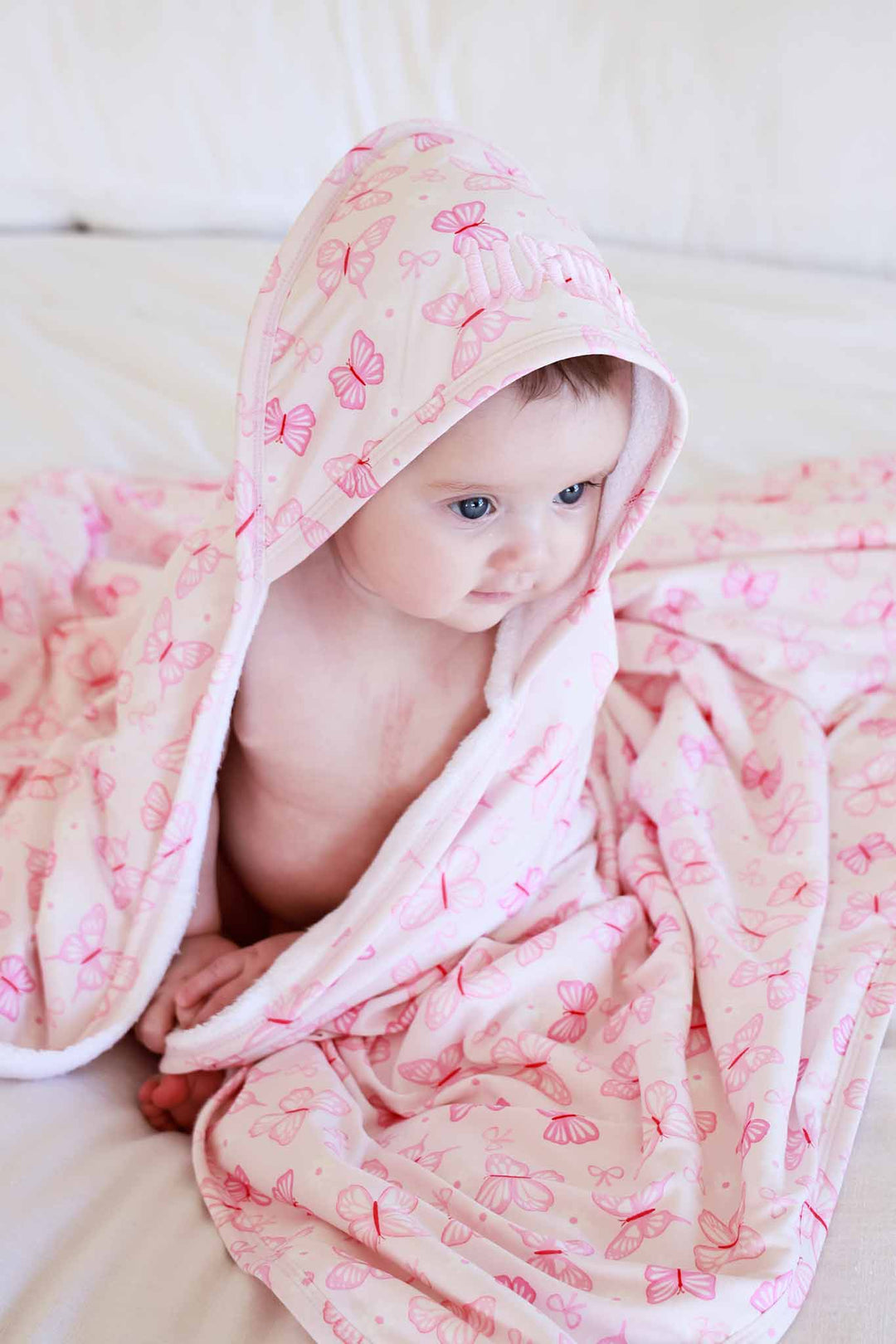 Embroidered Bamboo Hooded Towel in Blooming Butterflies print - baby sitting on white surface wrapped in the pink bamboo towel, with hood on and butterfly print visible