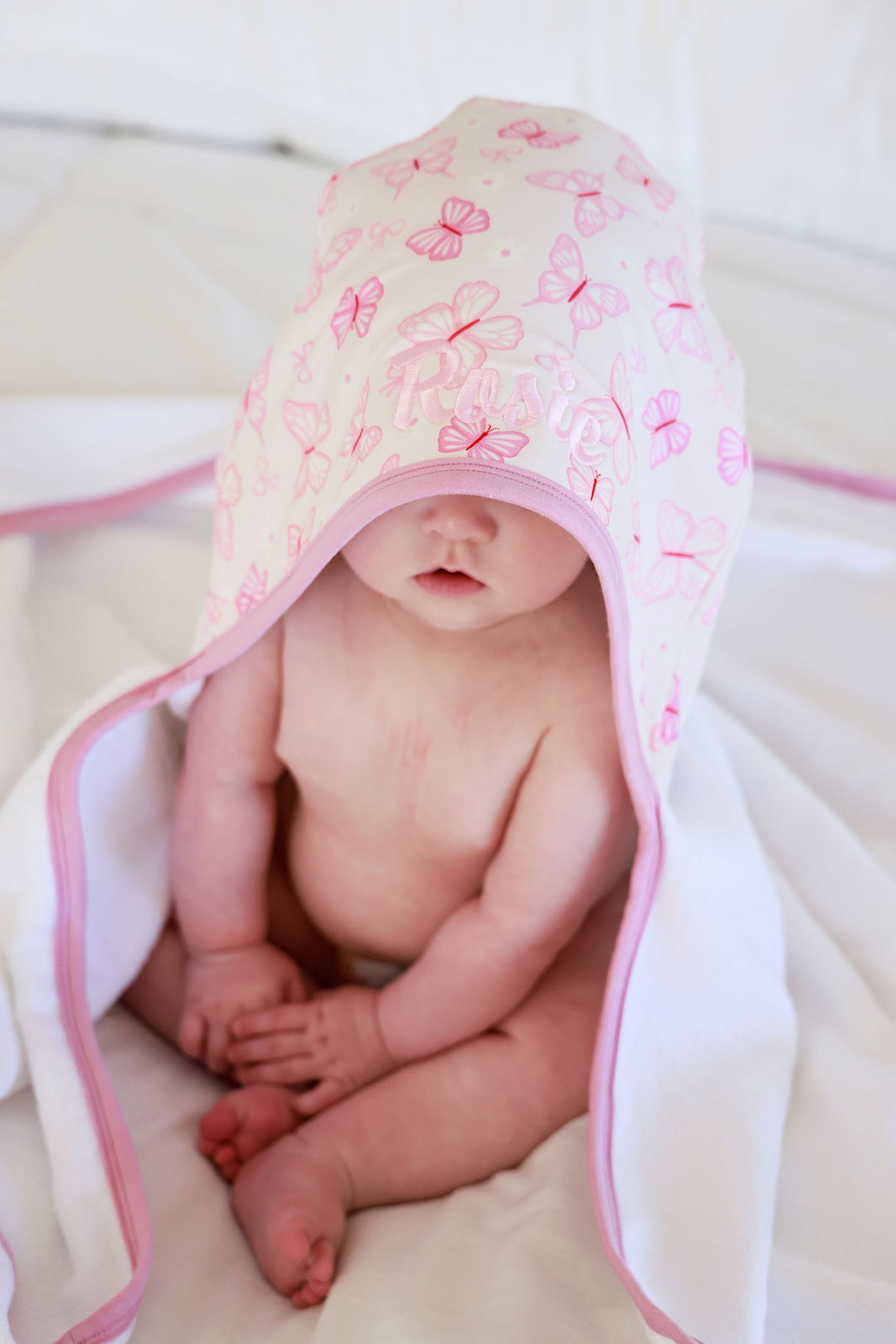 Embroidered Bamboo Hooded Towel in Blooming Butterflies print - baby lying on a white surface with the butterfly-print bamboo towel draped over them and the oversized hood pulled forward