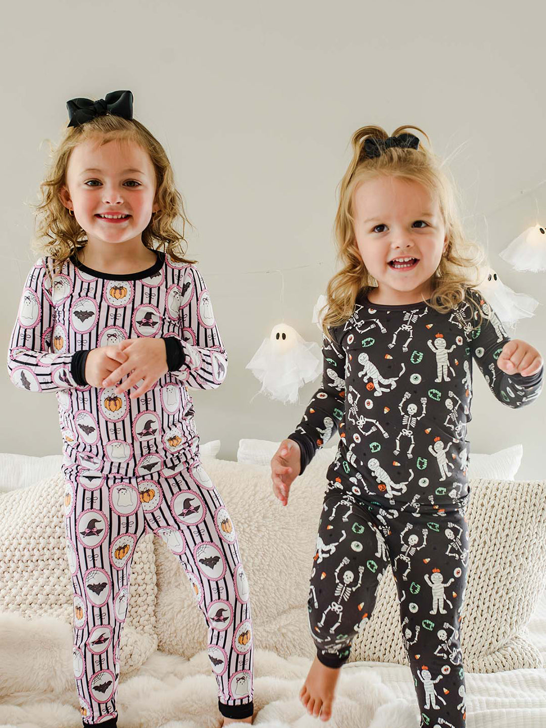 Young girls standing on a bed wearing  Caden Lane pink and black striped two-piece pajama sets with ghost, bat, and pumpkin cameo print, with a black bows in their hair and hanging ghost lights in the background.