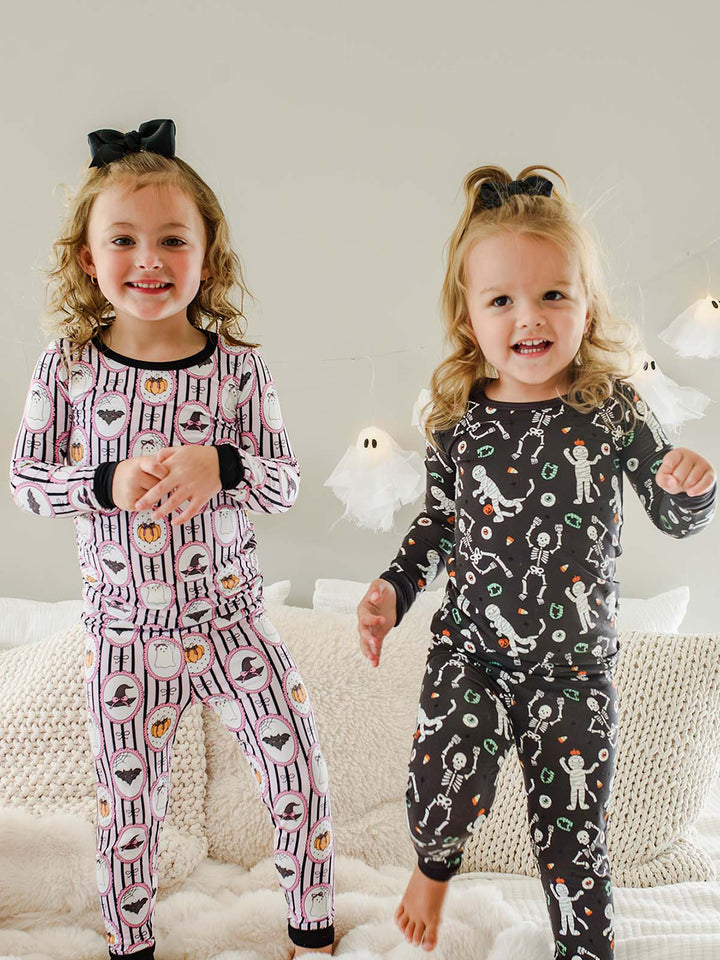Young girls standing on a bed wearing  Caden Lane pink and black striped two-piece pajama sets with ghost, bat, and pumpkin cameo print, with a black bows in their hair and hanging ghost lights in the background.