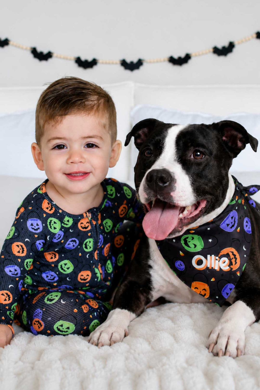 Toddler in candy-print pajamas sitting beside a dog wearing a matching personalized Halloween bandana with the name Ollie, bat garland on wall behind them