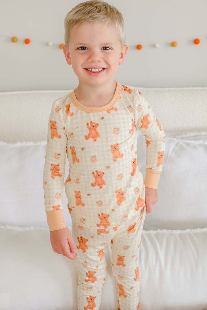 Laughing toddler boy wearing a Caden Lane honey bear print two-piece pajama set, standing on a bed