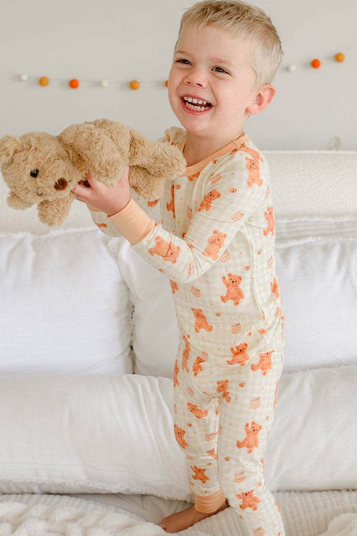 Laughing toddler boy wearing a Caden Lane honey bear print two-piece pajama set, standing on a bed holding up a plush teddy bear