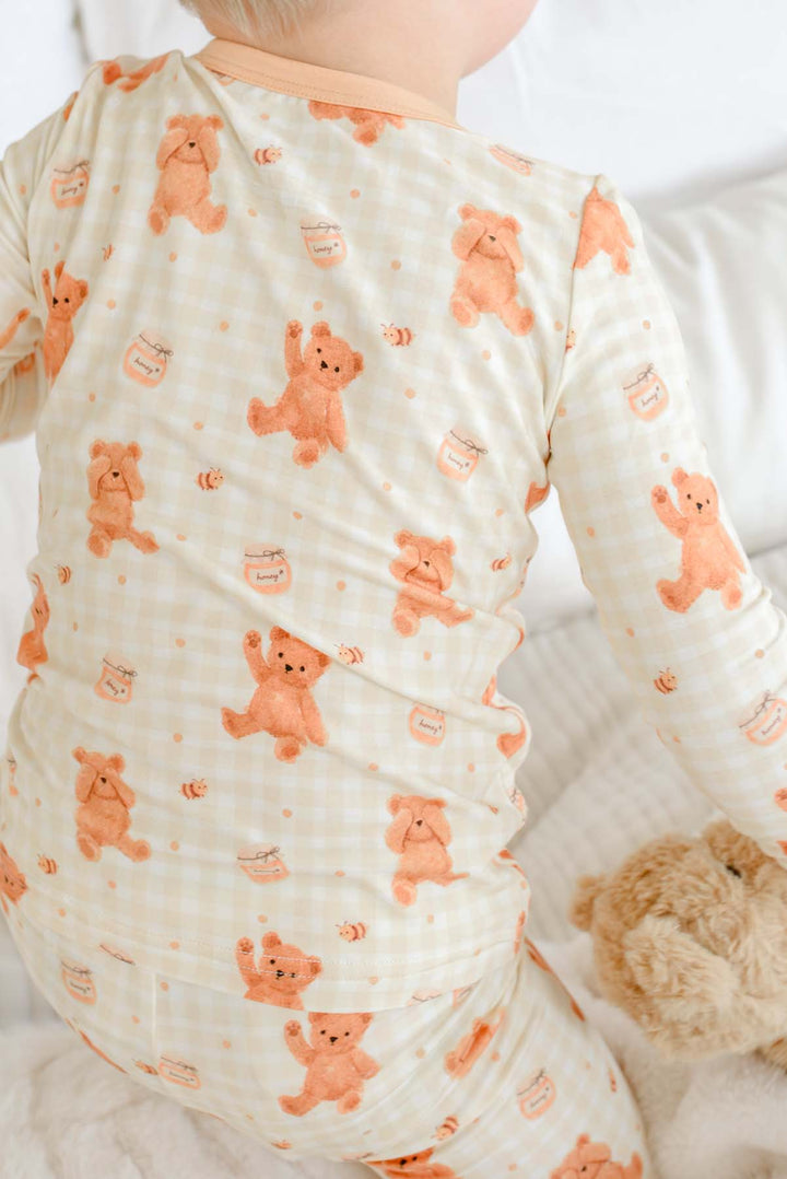 Toddler wearing a Caden Lane honey bear print two-piece pajama set, standing on a bed holding up a plush teddy bear