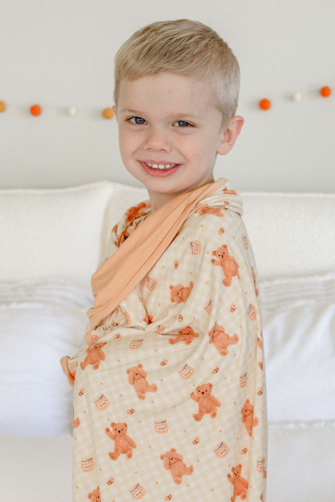Smiling boy wearing Caden Lane honey bear print pajamas, wrapped in a matching Caden Lane bamboo blanket while standing on a bed