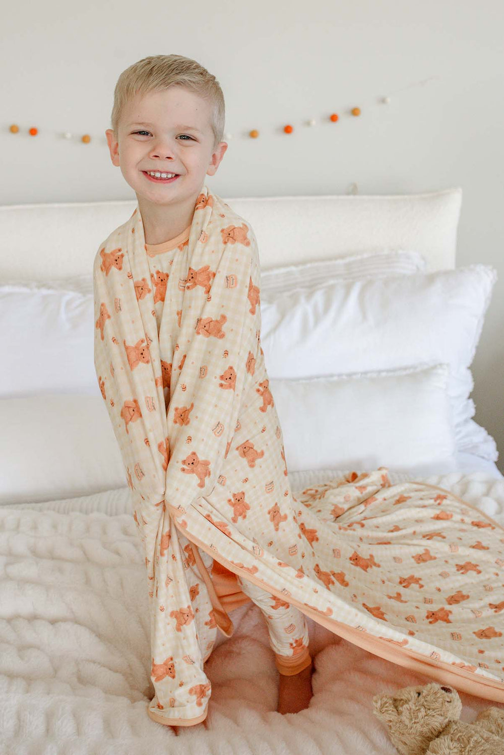 Smiling boy wearing Caden Lane honey bear print pajamas, wrapped in a matching Caden Lane bamboo blanket while standing on a bed