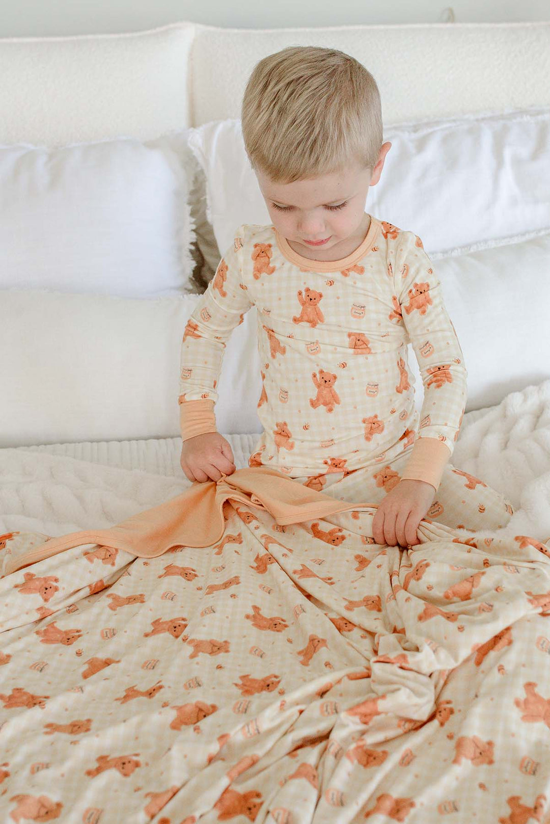 Smiling boy wearing Caden Lane honey bear print pajamas, wrapped in a matching Caden Lane bamboo blanket while kneeling on a bed