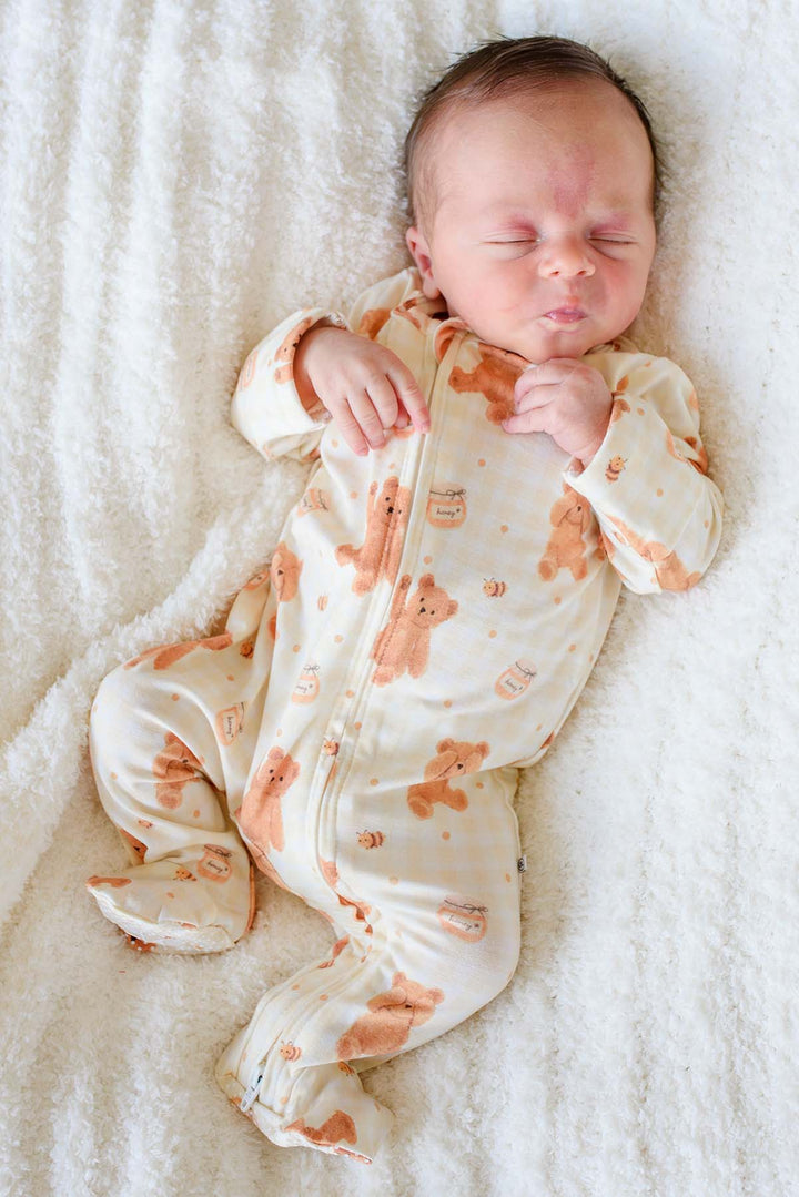 Newborn baby sleeping in a Caden Lane honey bear print footie pajama, lying on a fluffy white blanket