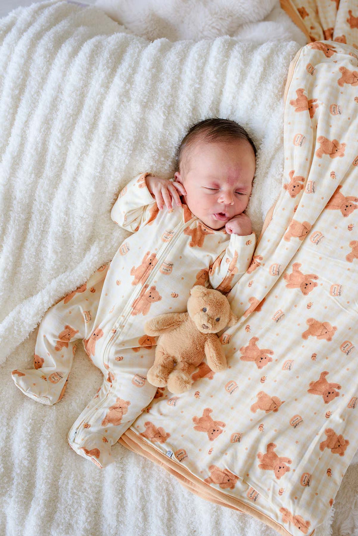 Newborn baby sleeping in a Caden Lane honey bear print footie pajama, lying on a fluffy white blanket next to a matching Caden Lane plush teddy bear and honey bear blanket