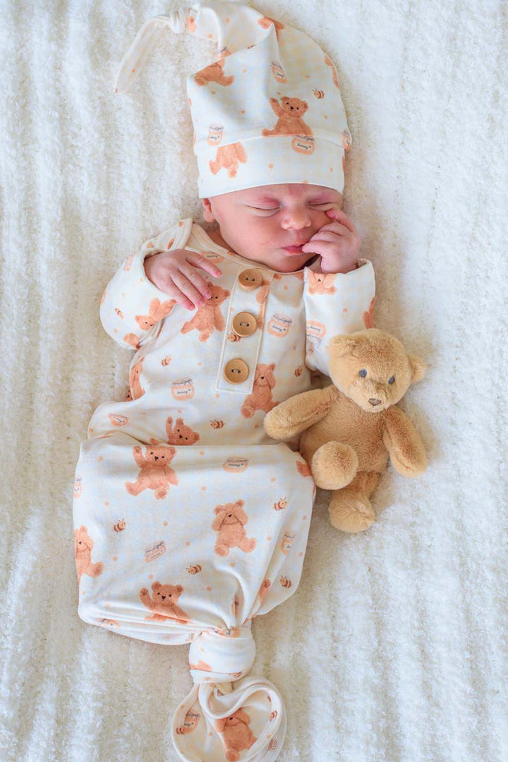 Sleeping newborn wearing a Caden Lane honey bear print knotted gown and matching sleeper cap, lying on a fluffy white blanket next to a plush teddy bear