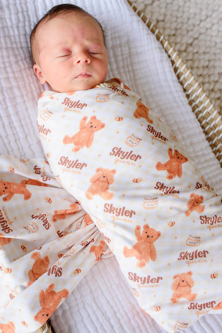Sleeping newborn swaddled in a Caden Lane personalized honey bear print swaddle reading 'Skyler Quinn,' lying in a woven bassinet 