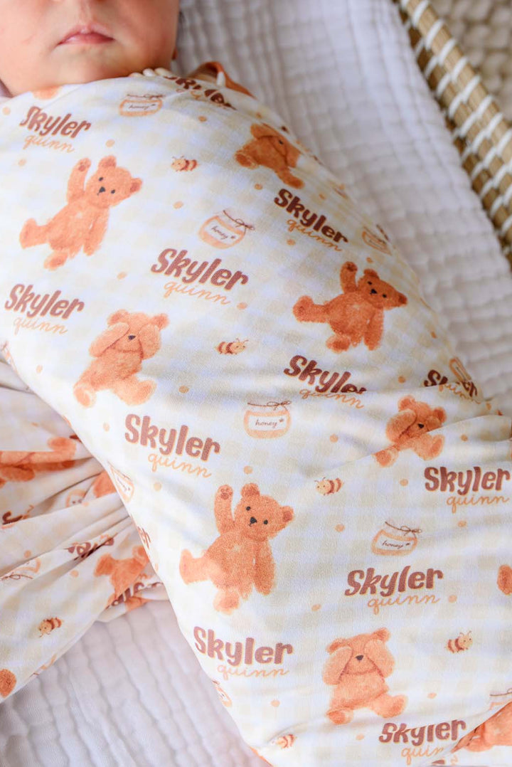 Newborn swaddled in a Caden Lane personalized honey bear print swaddle reading 'Skyler Quinn,' lying in a woven bassinet next to a plush teddy bear