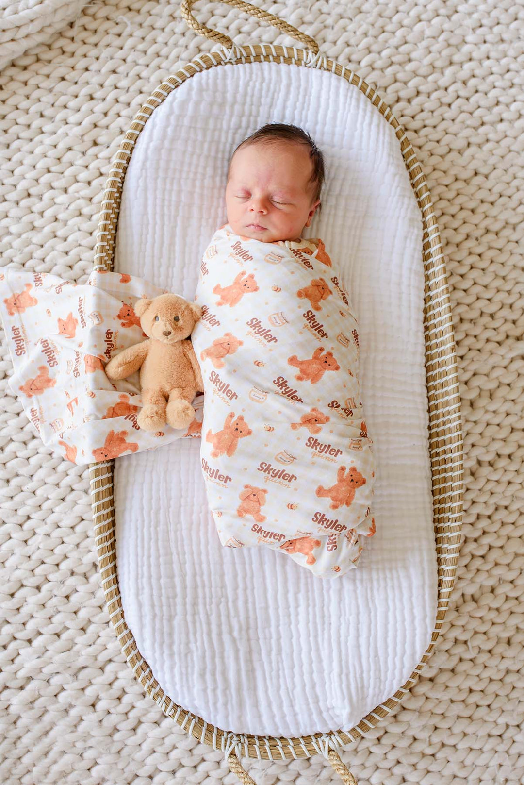Sleeping newborn swaddled in a Caden Lane personalized honey bear print swaddle reading 'Skyler Quinn,' lying in a woven bassinet next to a plush teddy bear