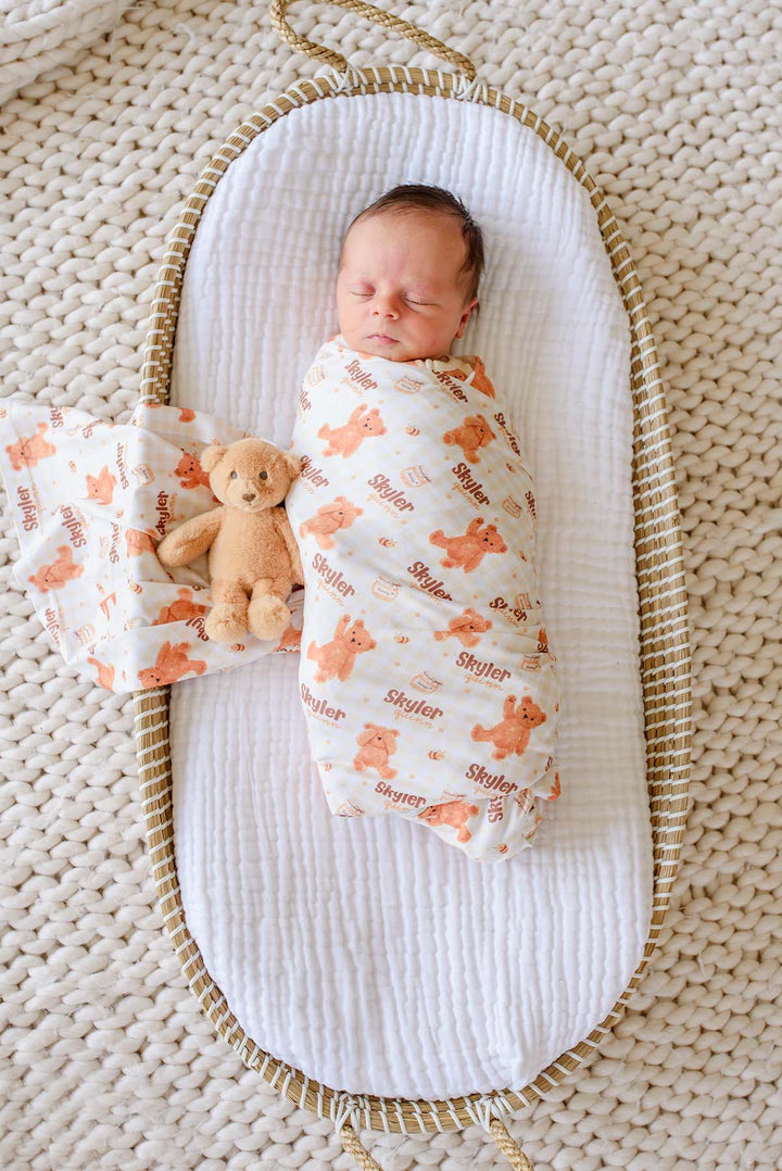 Sleeping newborn swaddled in a Caden Lane personalized honey bear print swaddle reading 'Skyler Quinn,' lying in a woven bassinet next to a plush teddy bear