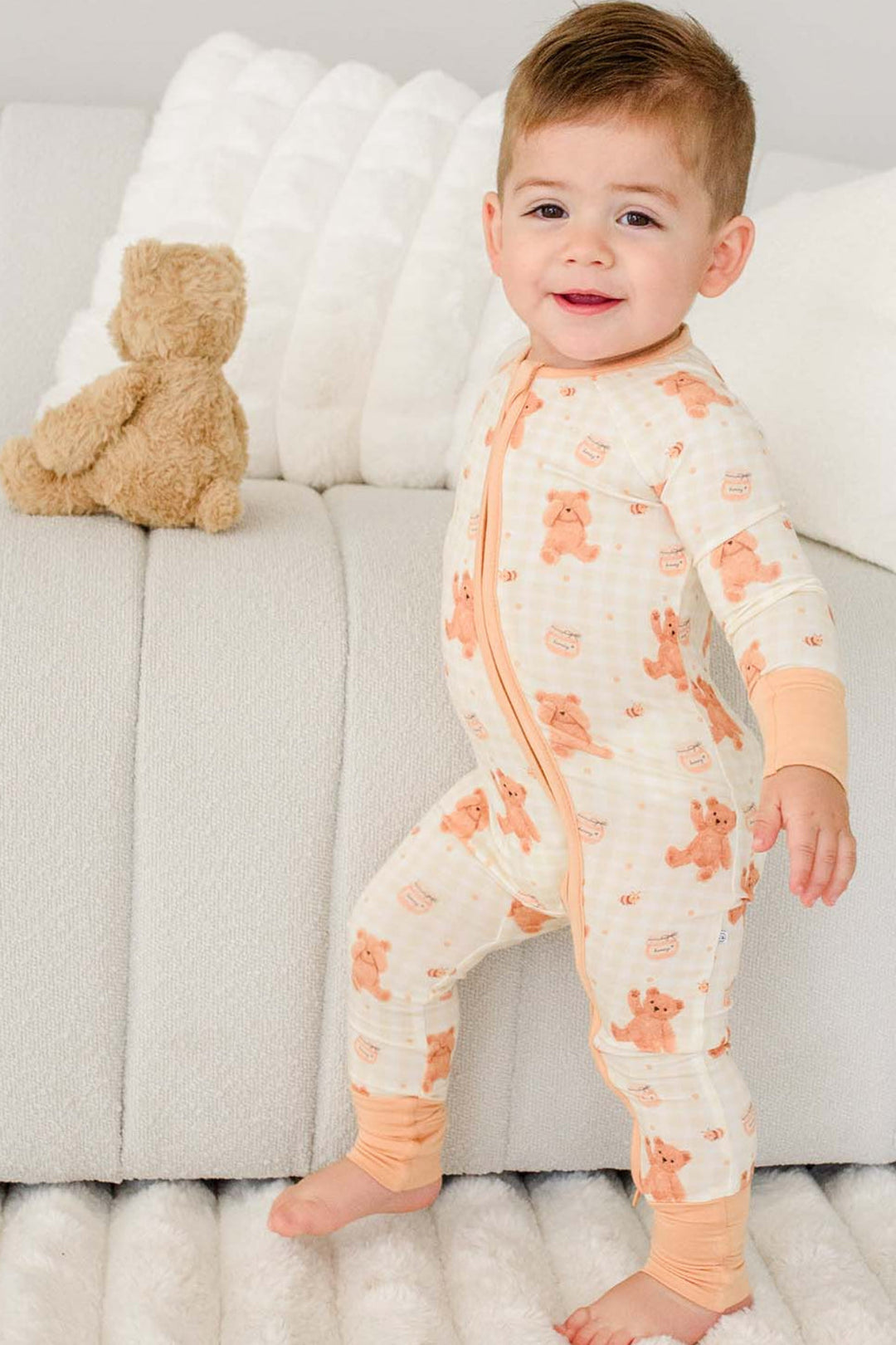 Smiling toddler standing barefoot on a couch wearing a Caden Lane honey bear print convertible romper, with a plush teddy bear on the cushion behind him