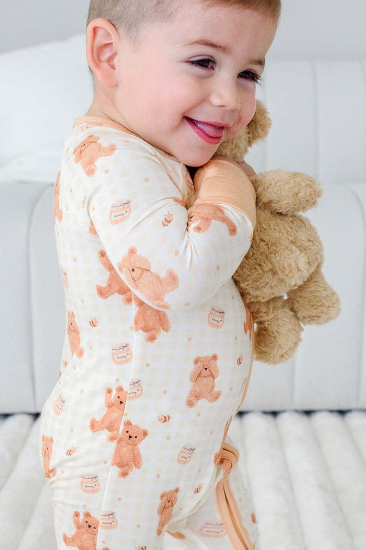 Smiling toddler standing wearing a Caden Lane honey bear print convertible romper, holding a plush teddy bear 