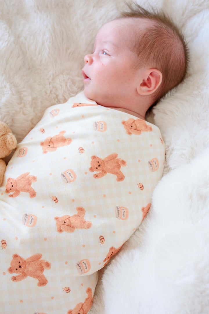 Newborn swaddled in a Caden Lane honey bear print swaddle blanket, lying on a fluffy white blanket next to a plush teddy bear