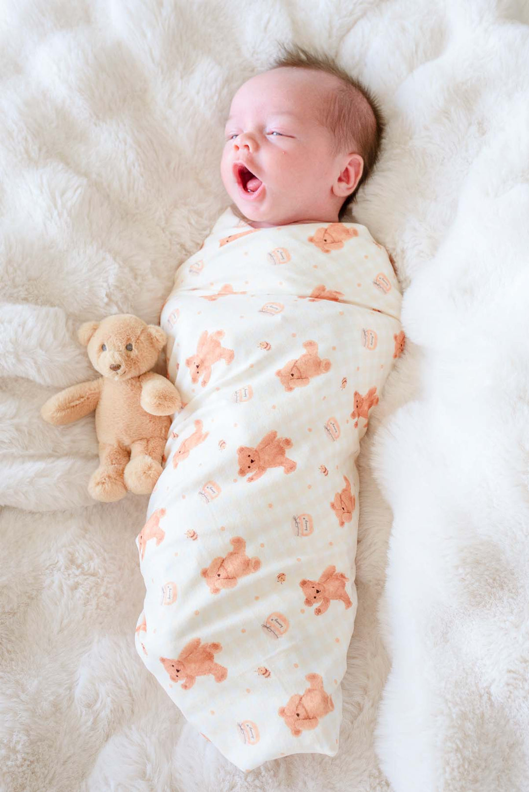 Yawning newborn swaddled in a Caden Lane honey bear print swaddle blanket, lying on a fluffy white blanket next to a plush teddy bear
