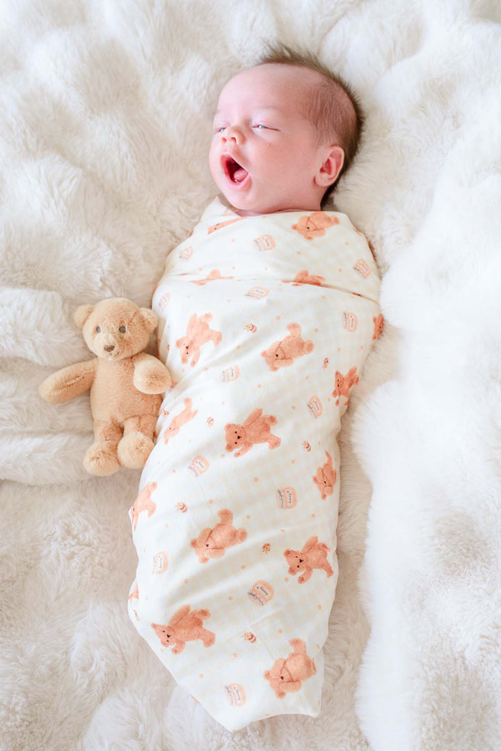 Yawning newborn swaddled in a Caden Lane honey bear print swaddle blanket, lying on a fluffy white blanket next to a plush teddy bear