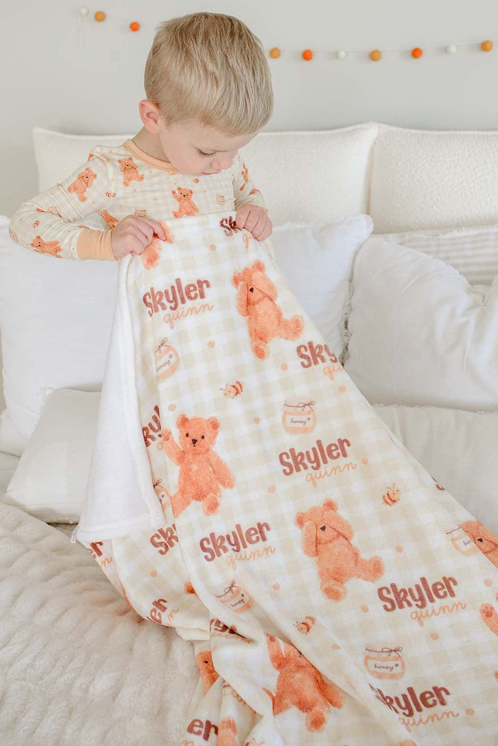 Boy in Caden Lane honey bear print pajamas standing on a bed, holding a personalized Caden Lane blanket printed with 'Skyler Quinn'
