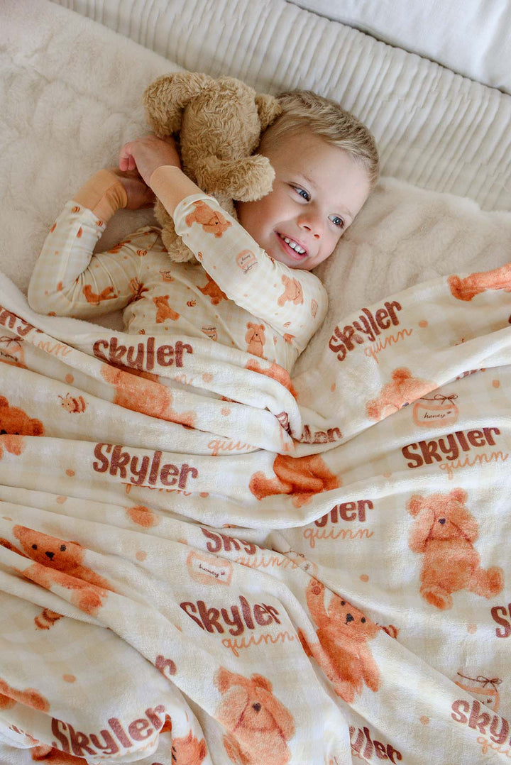Smiling boy in Caden Lane honey bear print pajamas laying on a bed, holding a plush teddy bear with a personalized Caden Lane blanket printed with 'Skyler Quinn'