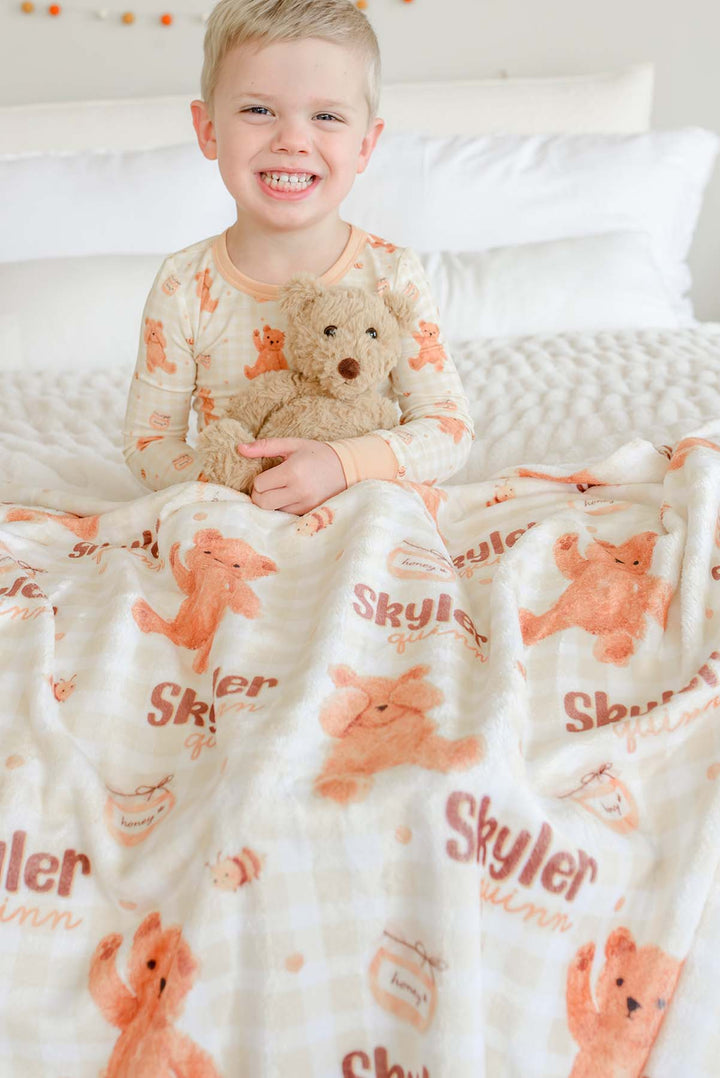 Smiling boy in Caden Lane honey bear print pajamas sitting on a bed, holding a plush teddy bear beside a personalized Caden Lane blanket printed with 'Skyler Quinn'