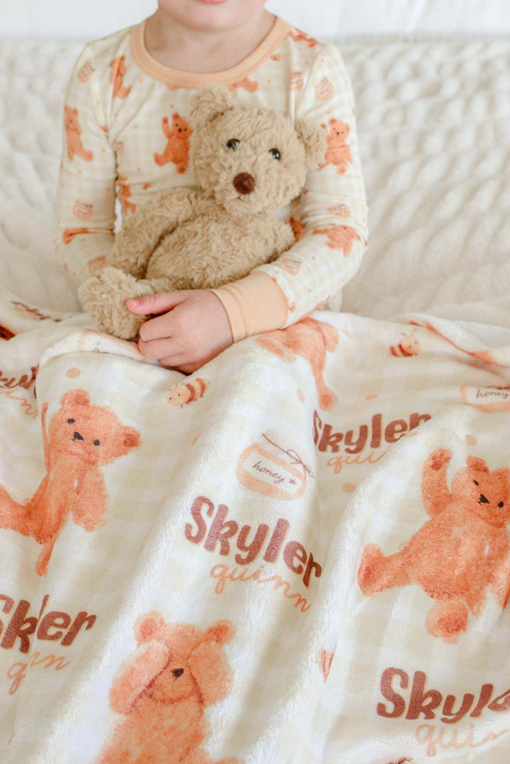 Boy in Caden Lane honey bear print pajamas sitting on a bed, holding a plush teddy bear beside a personalized Caden Lane blanket printed with 'Skyler Quinn'