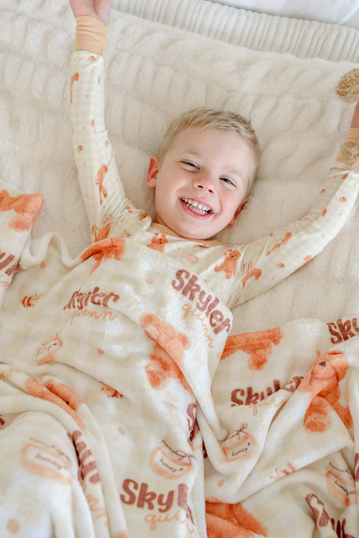 Smiling boy in Caden Lane honey bear print pajamas laying on a bed, holding a plush teddy bear with a personalized Caden Lane blanket printed with 'Skyler Quinn'