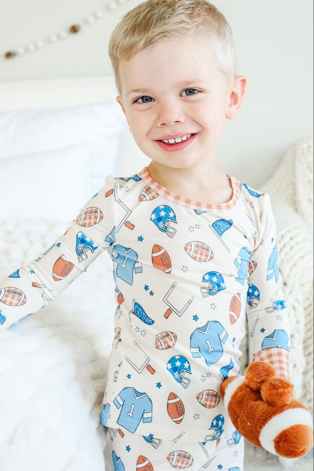 Little boy standing next to a bed and wearing the Caden Lane blue huddles and cuddles two piece pajama set while holding a football stuffy. 