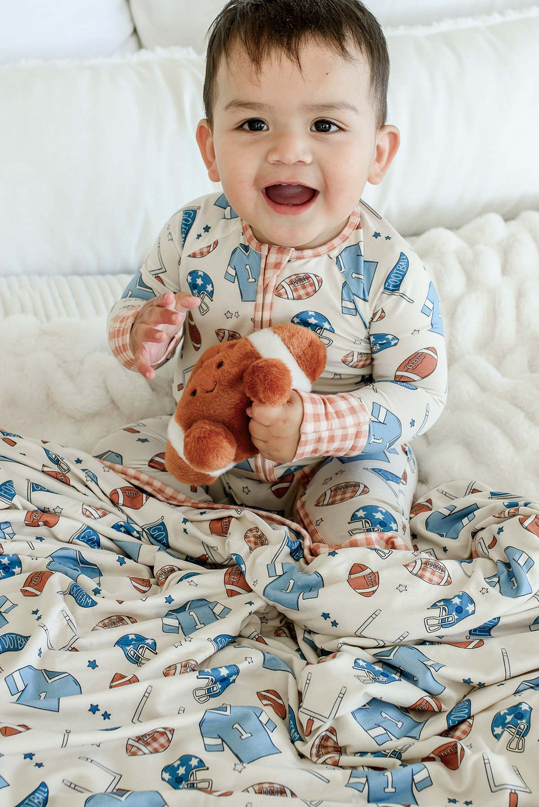 Baby sitting holding a stuffed smiling football on a white fuzzy blanket, wearing a Caden Lane 'Huddles and Cuddles' blue football-print outfit with orange gingham trim, with a matching bamboo blanket