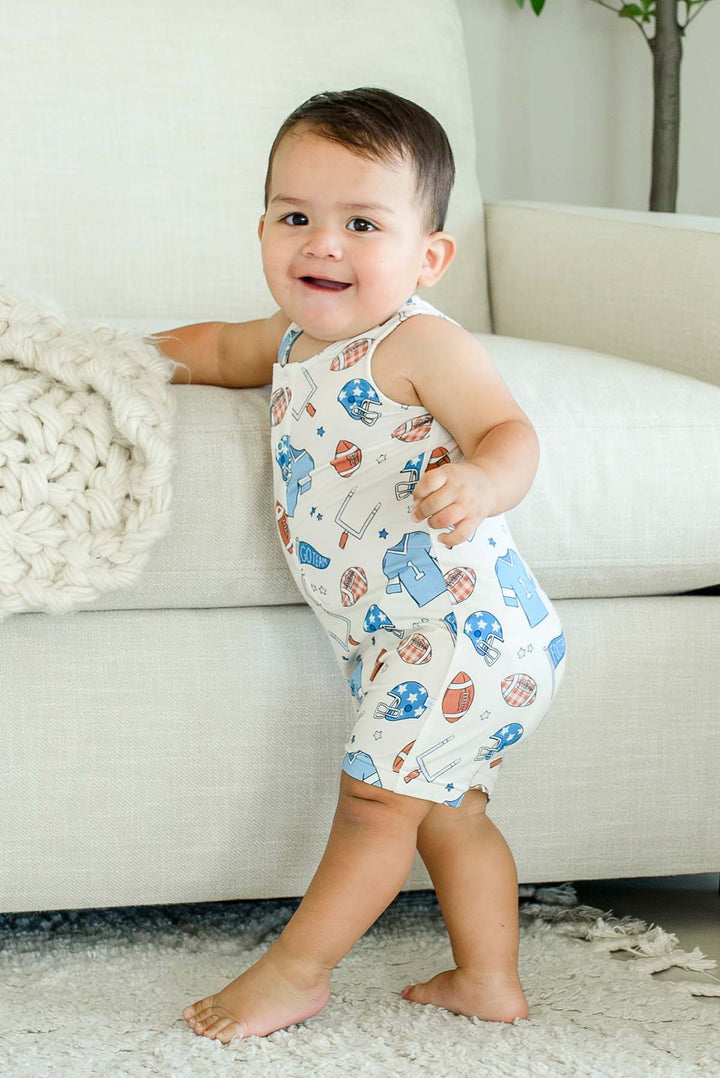 Wide shot of a smiling baby standing barefoot on a rug, wearing a Caden Lane 'Huddles and Cuddles' blue football-print sleeveless romper, leaning on a cream couch