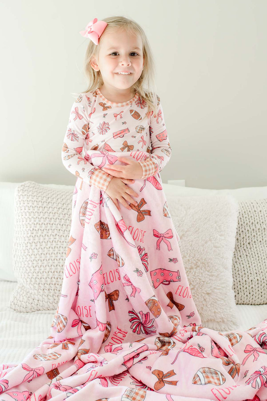Smiling young girl standing on bed, with a pink hair bow, wrapped in a personalized Caden Lane 'Huddles and Cuddles' pink football and cheerleading print blanket