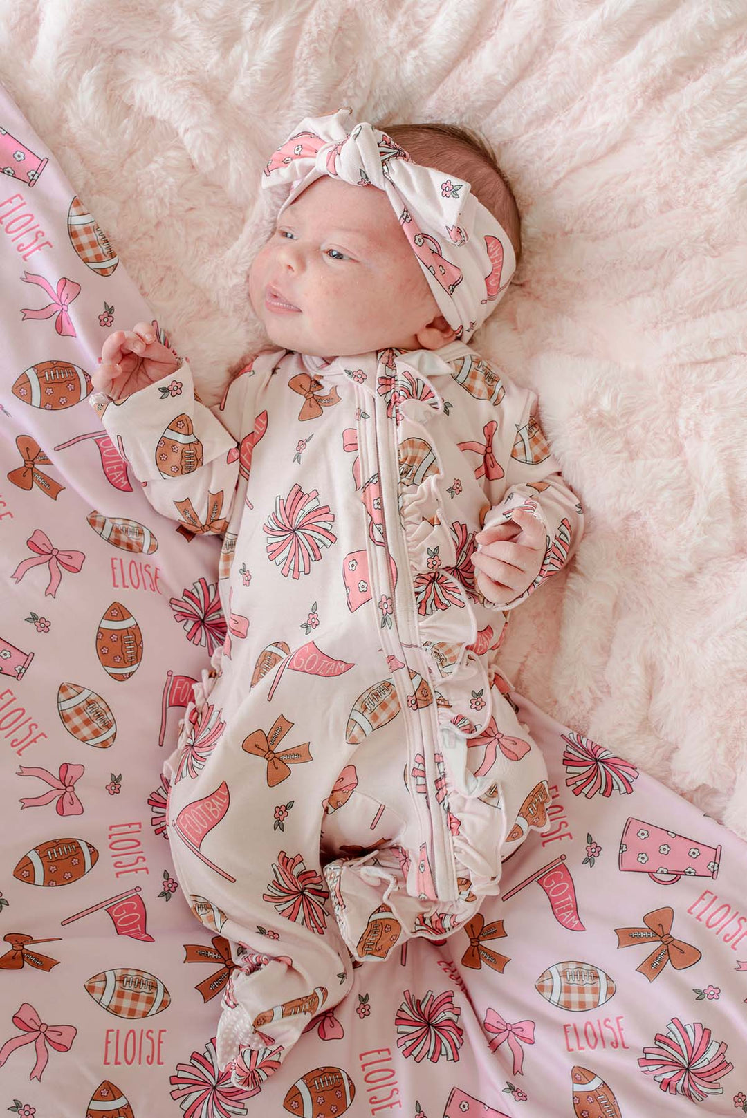 A baby lying on a fluffy cream blanket, wearing a cream zip-front footed sleeper with a matching headband and a pink patterned blanket underneath. The pattern features pink bows, brown footballs, and the word 'ELOISE'.