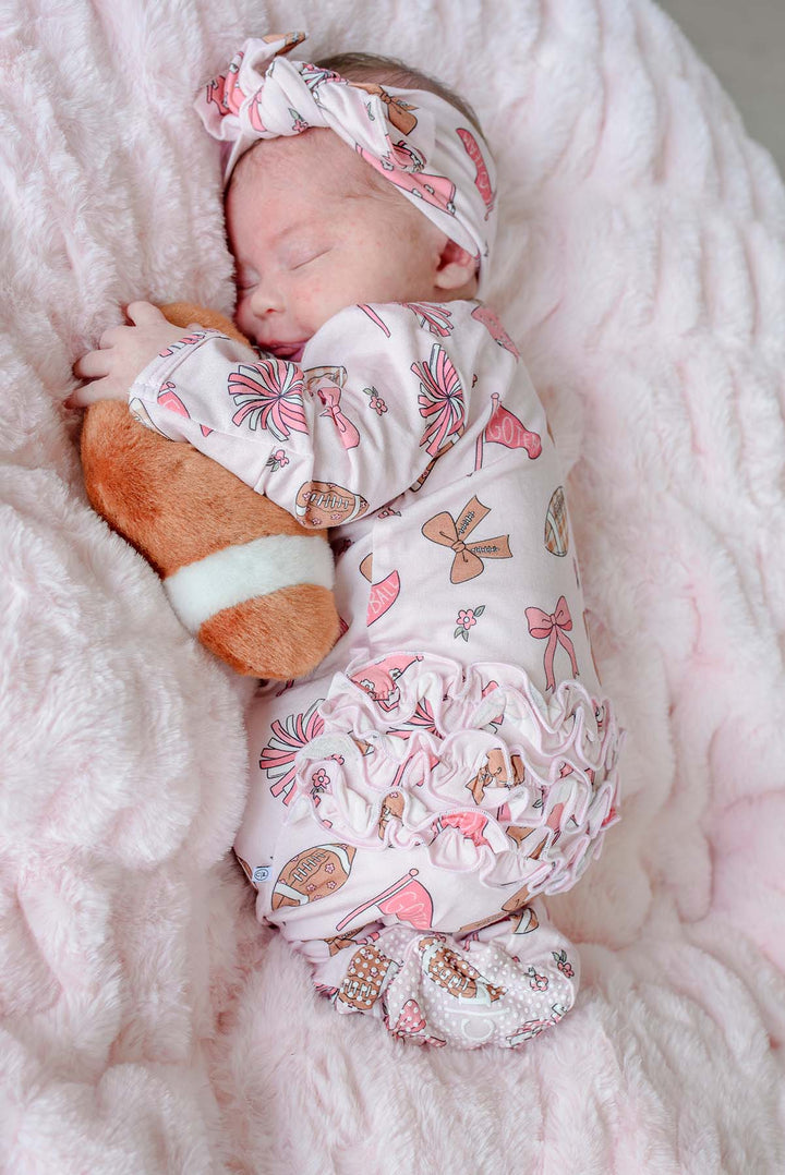 A sleeping newborn baby wearing a light pink ruffled romper and matching headband with a pink, brown, and white pattern, lying on a fluffy pink blanket and holding a plush toy.