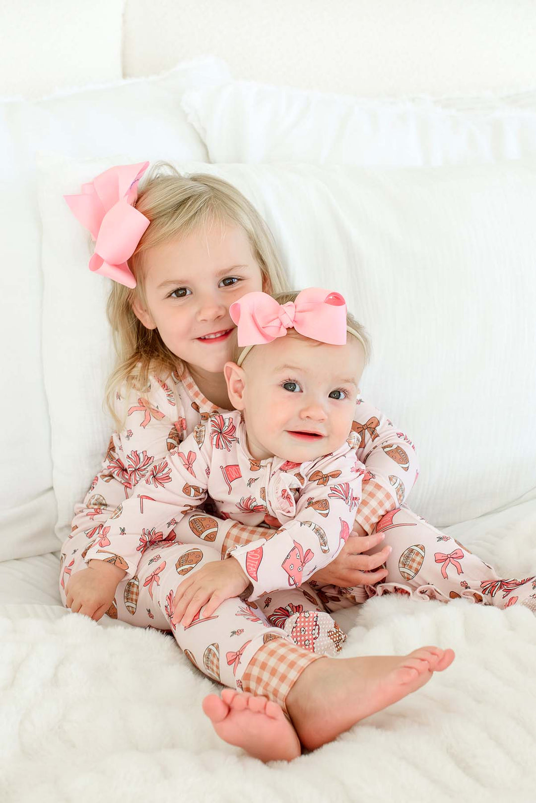 Two young girls sitting together on a white bed, wearing matching pink and brown patterned pajamas and large pink bows in their hair.