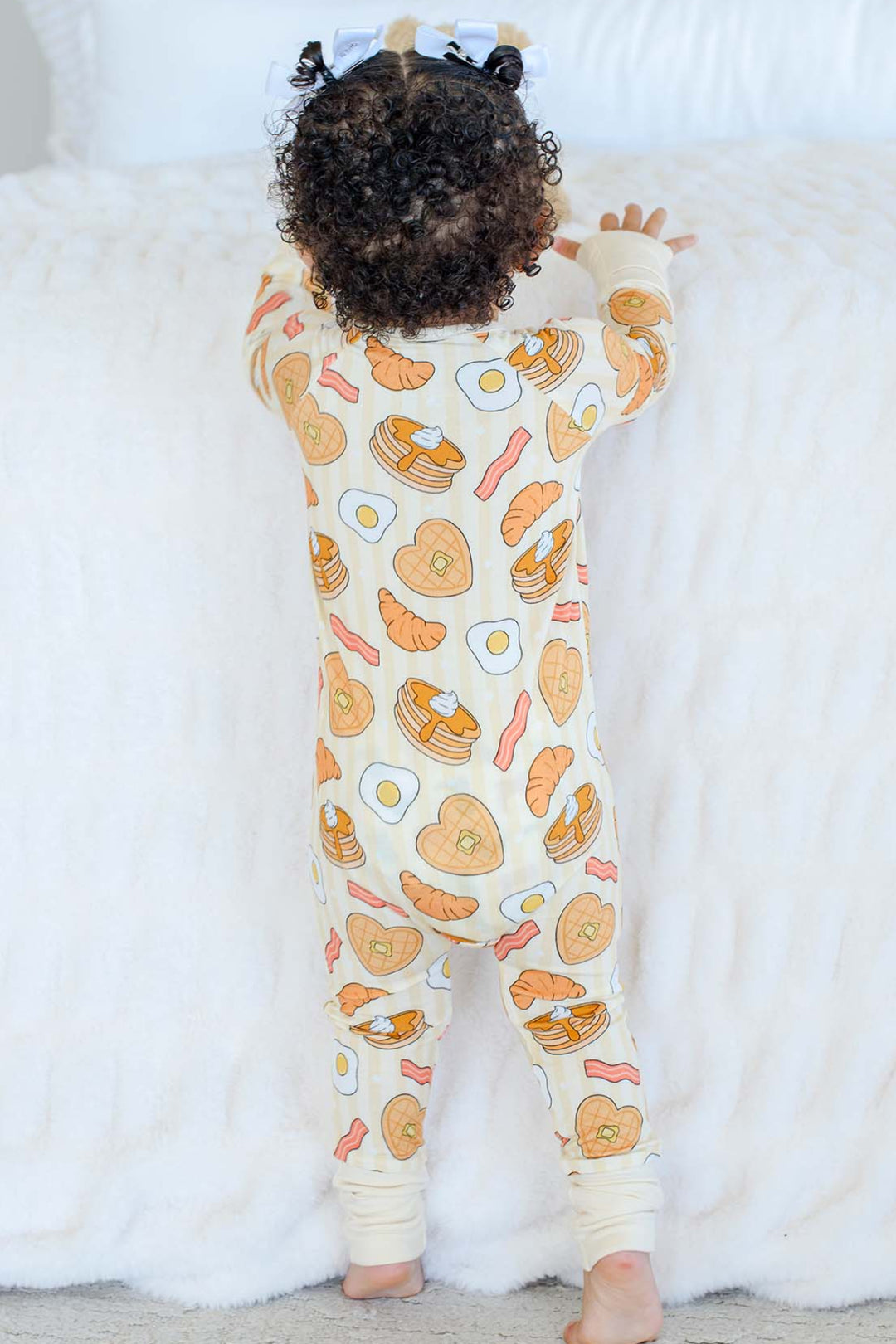 Back view of toddler standing in Caden Lane zip footie pajama with breakfast-themed print, hair in white bows