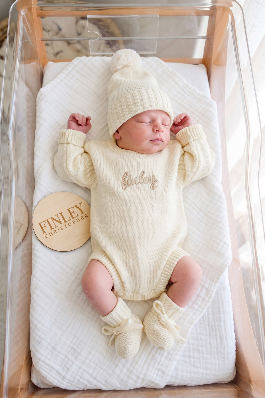 Sleeping newborn in a bassinet wearing Caden Lane ivory personalized knit sweater romper "Finley," matching pom-pom hat, and tie booties, beside a wooden name sign
