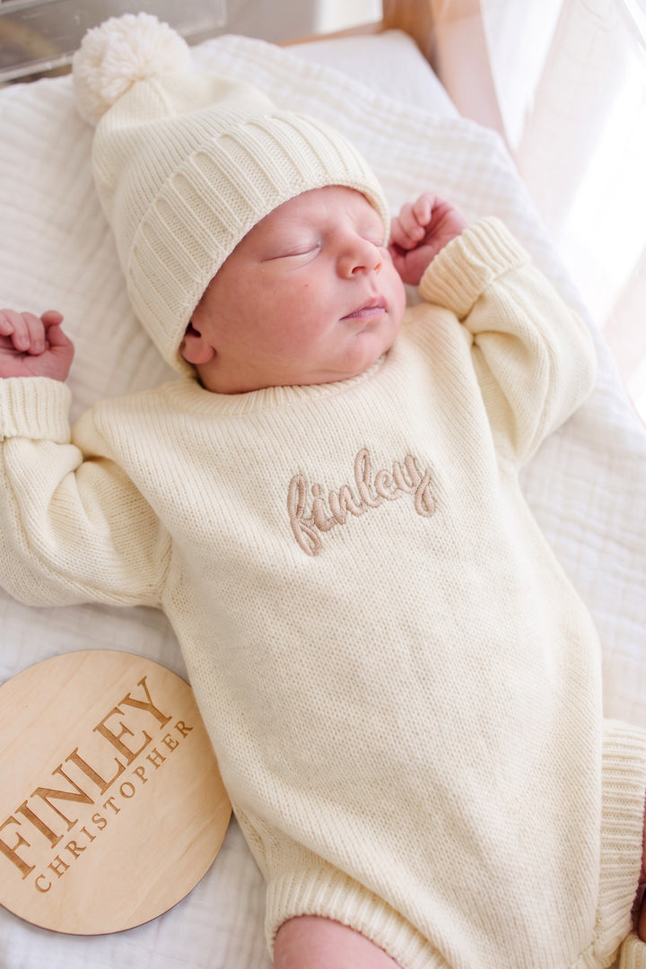 Close-up of sleeping baby wearing Caden Lane ivory knit romper with "Finley" script name embroidery and matching pom-pom beanie