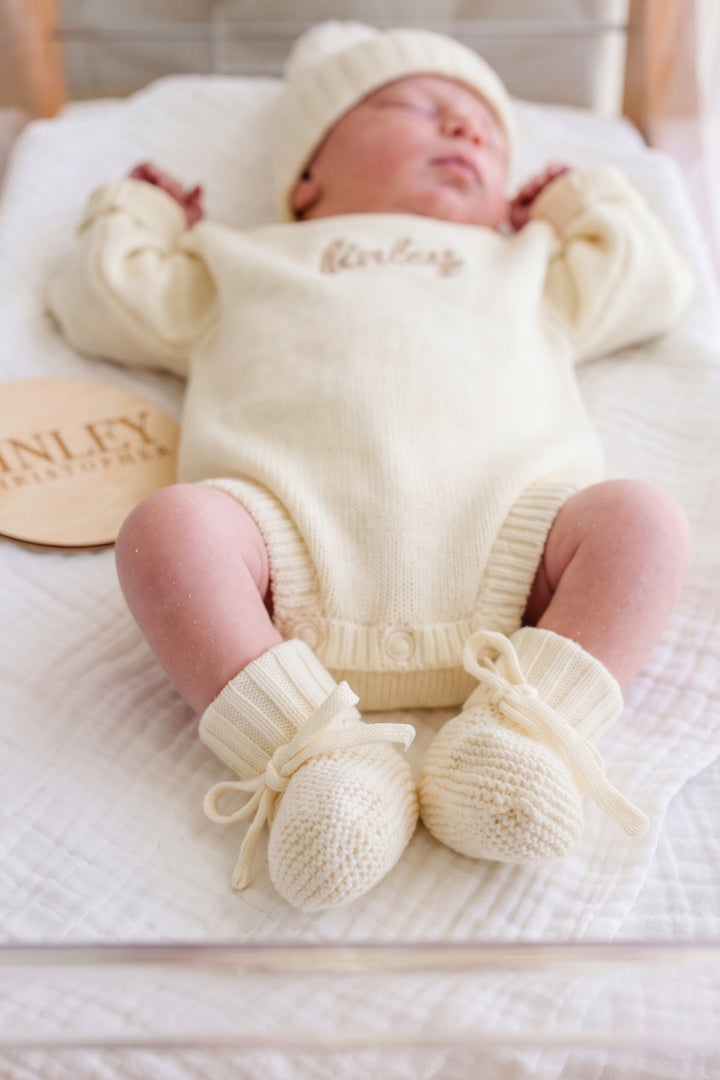 Newborn's legs and feet in Caden Lane ivory knit tie booties, wearing matching personalized "Finley" romper beside a wooden name sign