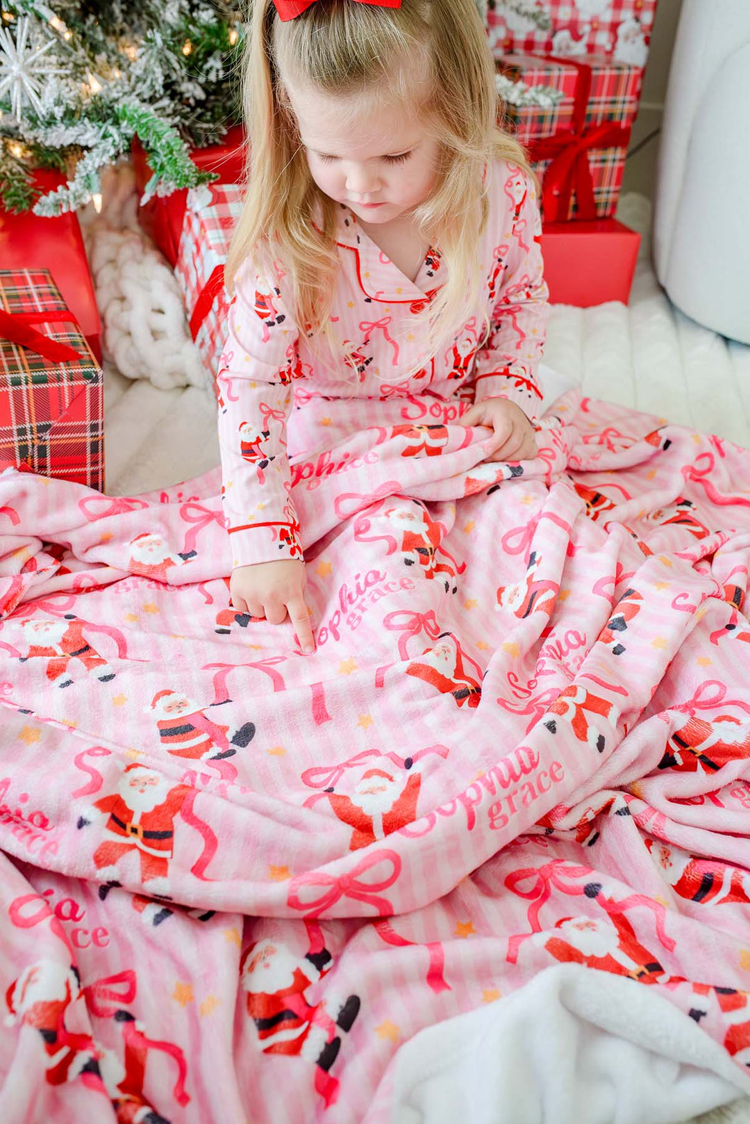 A young girl sits on the floor wrapped in a pink Christmas-themed blanket and wearing matching pajamas with Santa Claus prints, surrounded by wrapped gifts and a decorated tree.