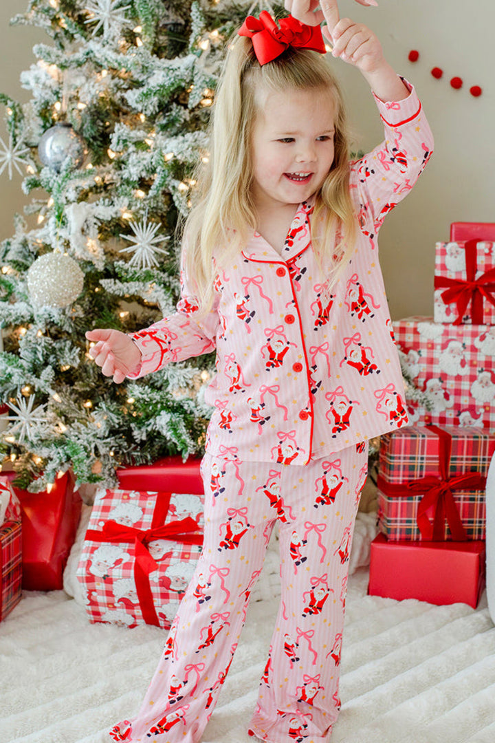 Girl wearing the Caden Lane Jolly Ribbons Girls' Christmas Two-Piece Flare Lounge Set featuring festive Santas and ribbon bows while twirling beside a decorated Christmas tree.