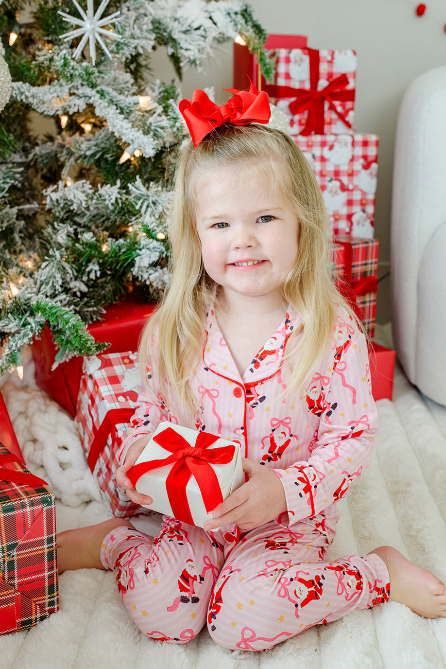 Girl wearing the Caden Lane Jolly Ribbons Christmas Bamboo Two-Piece Pajama Set, holding a wrapped Christmas gift beside a decorated holiday tree.