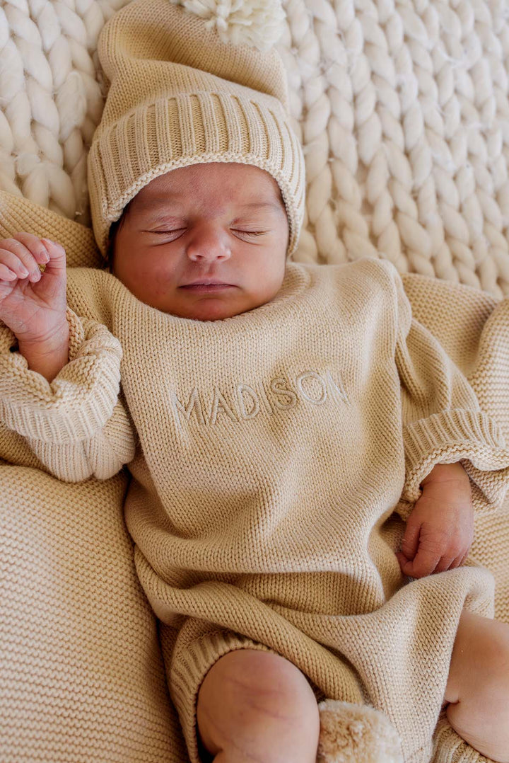 Baby's hand and feet in Caden Lane beige knit bubble romper with matching pom-pom booties, personalized name embroidery