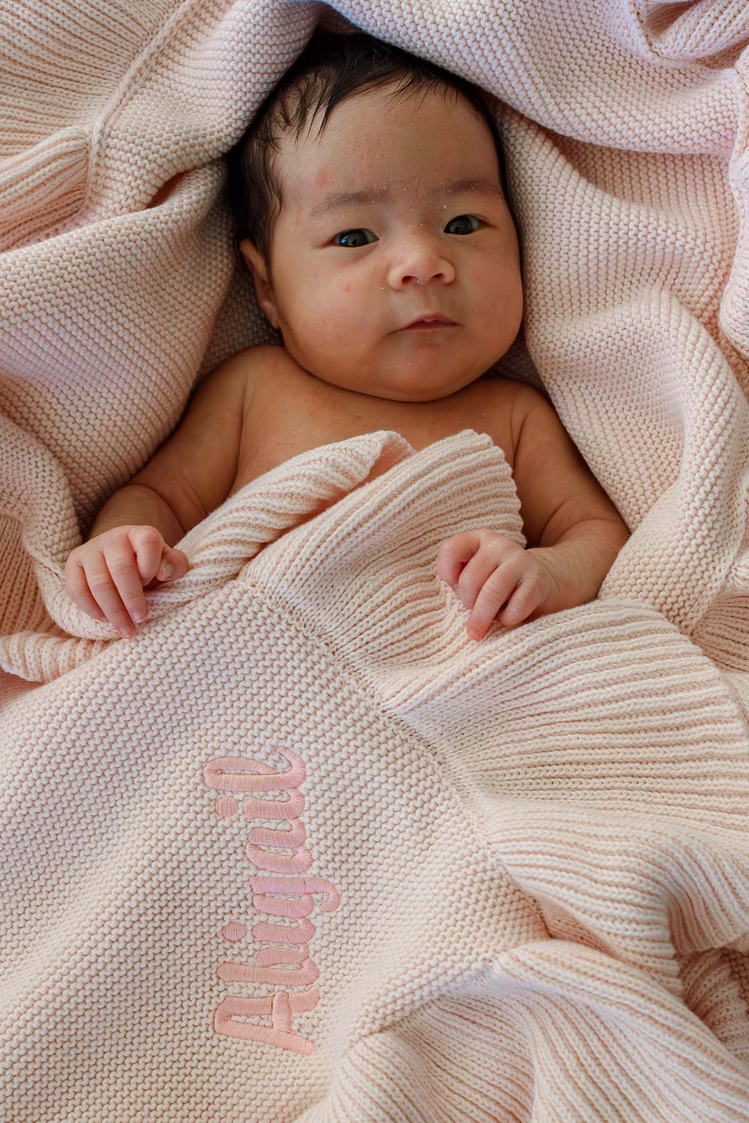 A baby wrapped in a soft cream-colored knit blanket with the name 'Abigail' embroidered in pink script, lying on a matching textured blanket.